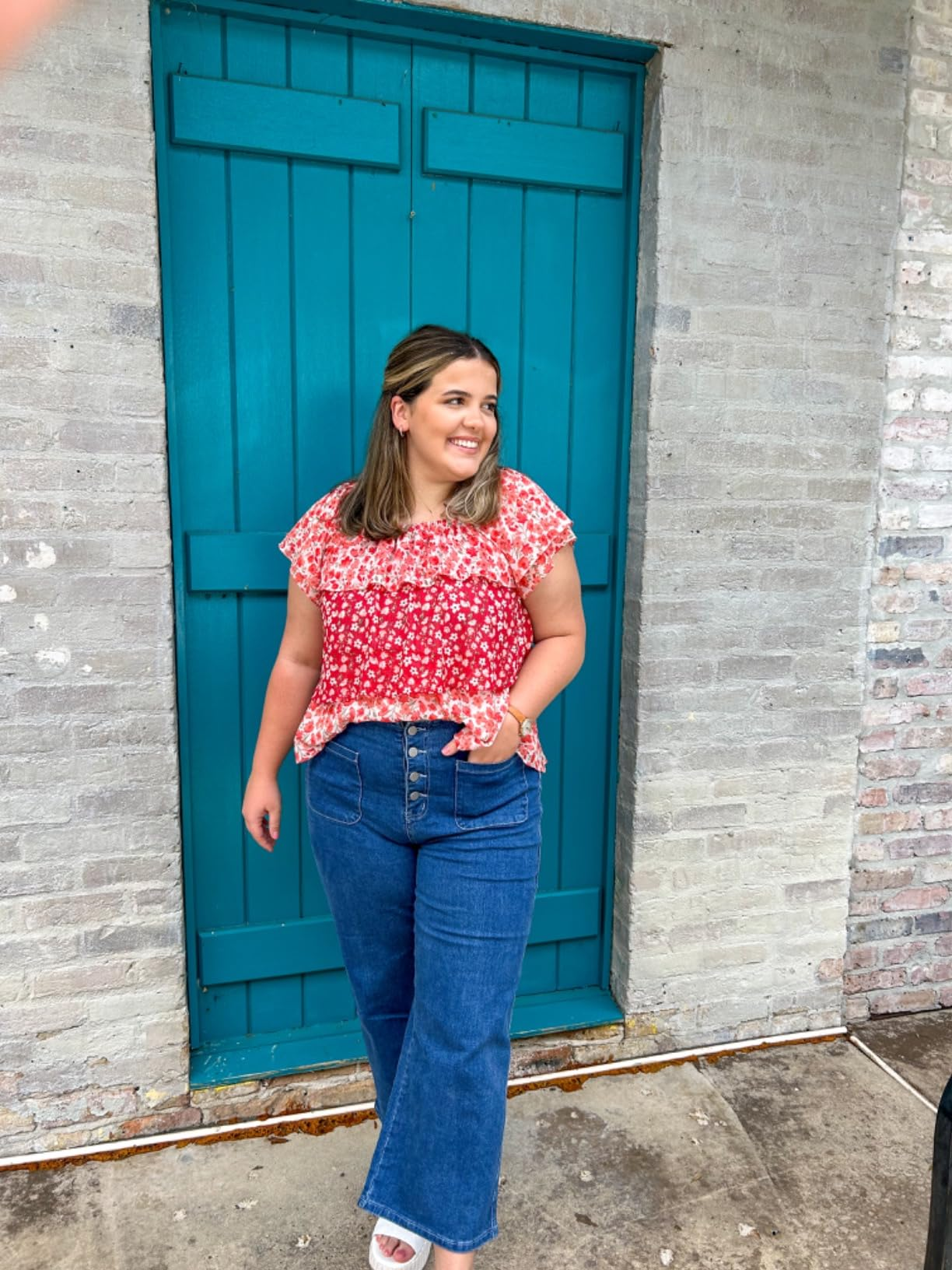 Person wearing a floral blouse and jeans poses in front of a brick wall with a wooden door