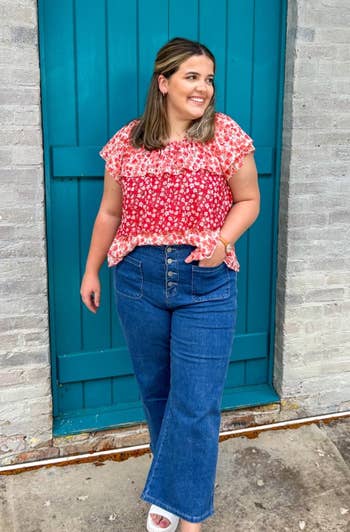 Person wearing a floral blouse and jeans poses in front of a brick wall with a wooden door