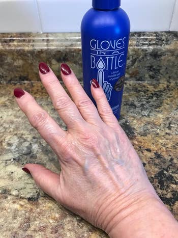 A person's hand with painted nails near a bottle of Gloves In A Bottle lotion, positioned on a countertop