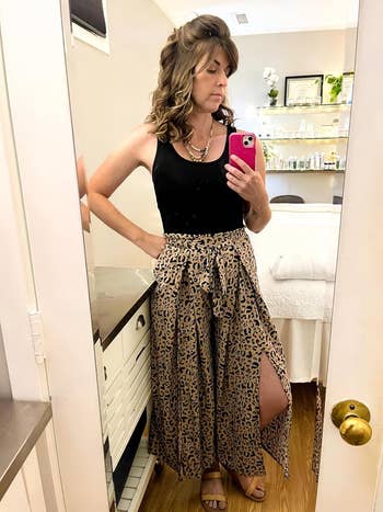 Person in a black tank top and leopard print skirt looks in the mirror, taking a selfie inside a room