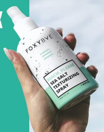 A hand holds a bottle of FoxyBae Beach, Please Sea Salt Texturizing Spray. The label mentions it includes biotin