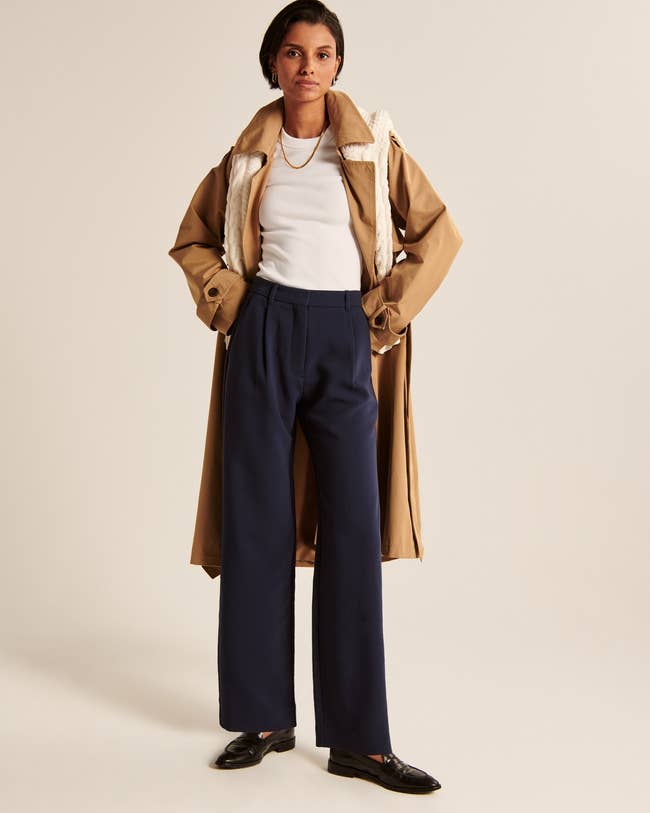 Model in a trench coat over a white top, navy trousers, and loafers