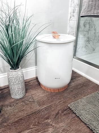 A towel warming machine with a wooden base and lid next to a decorative plant in a patterned vase, in a bathroom setting