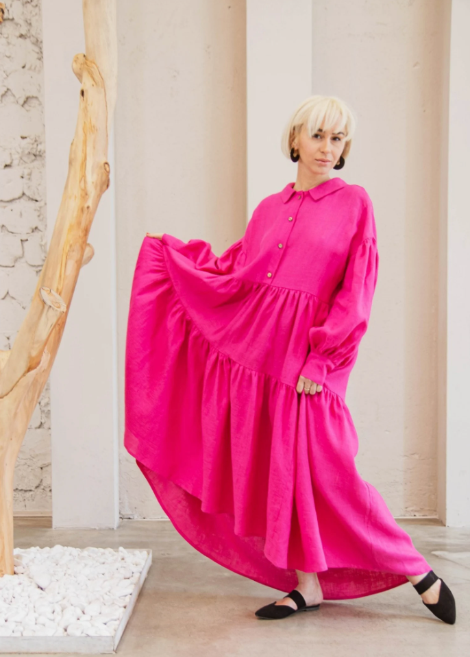 Model wearing a tiered hot pink maxi dress with a button down top portion and collar in black flats while lifting the skirt up