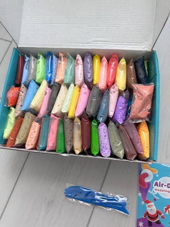 Box of colorful modeling clay packets with 2 bluish tools beside it connected a woody surface