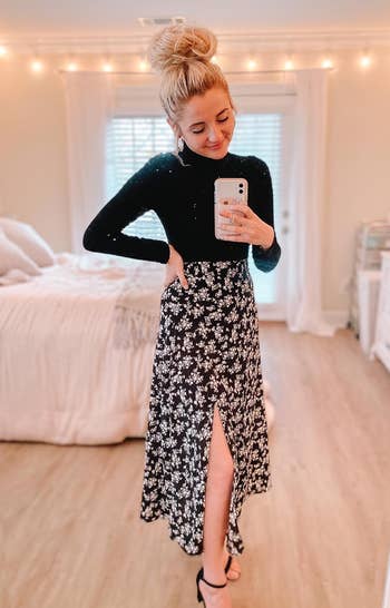 reviewer in a long sleeve black top and floral skirt with a high slit, taking a selfie in a bedroom