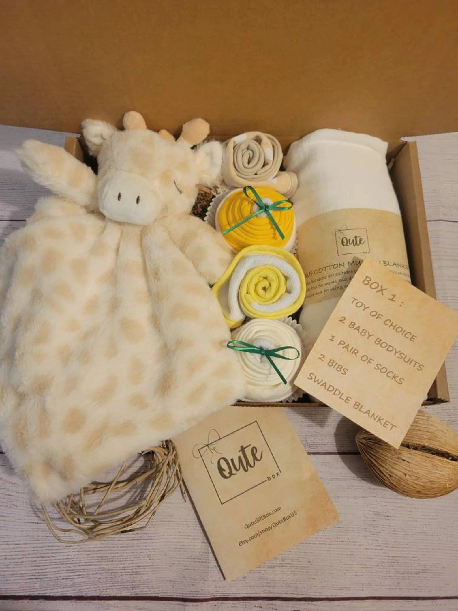 Brown box filled with plush giraffe toy, white rolled up blanket, and yellow, brown, and white rolled baby clothes and bibs