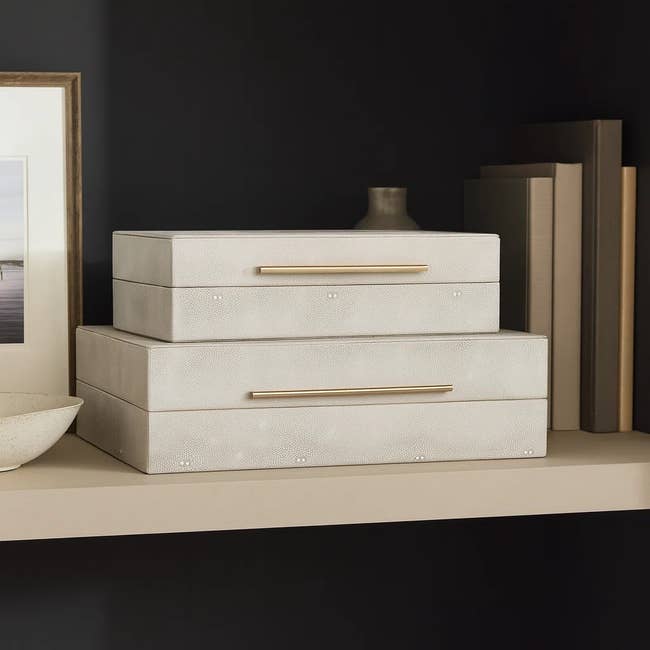 Two stacked jewelry boxes with gold handles on a shelf, surrounded by framed art, books, and a ceramic vase