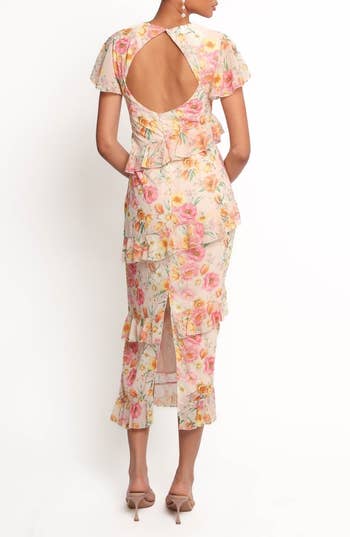 A person wearing a floral ruffle dress with open back and midi length, posing in heels, seen from behind