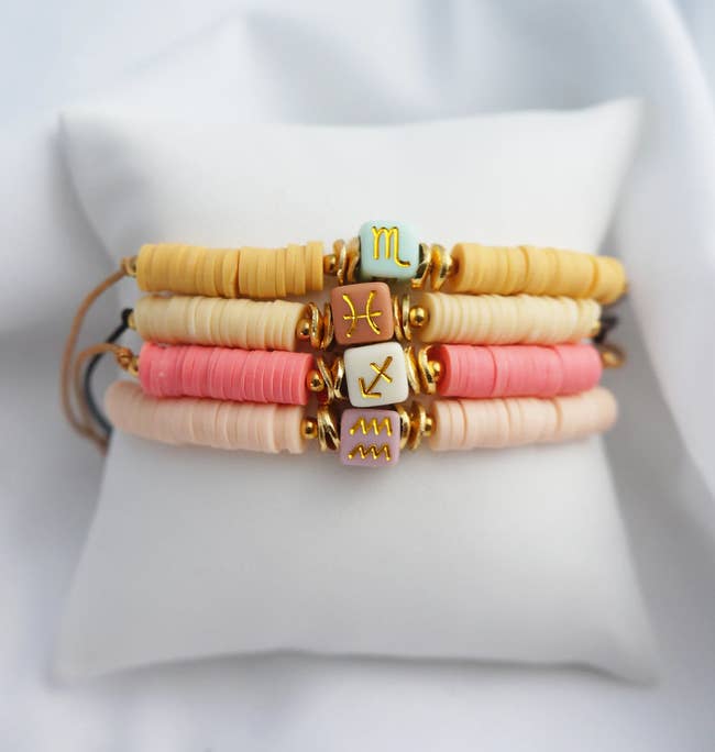 Four beaded bracelets in yellow, tan, pink, and light pink with zodiac sign clay beads in blue, red, white, and purple wrapped around a white pillow
