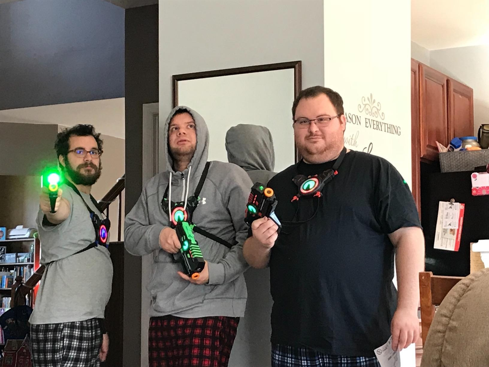 Three radical   basal   indoors wearing laser tag gear, holding laser guns. Casual attire includes pajamas and hoodies. They look  acceptable   for a game