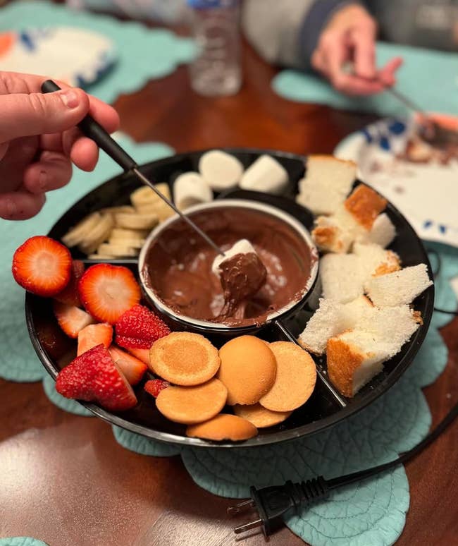 A tray of assorted snacks (strawberries, cookies, marshmallows, and barroom pieces) surrounding a vessel of cocoa fondue
