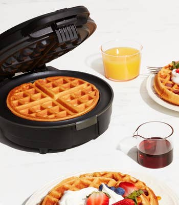 Waffle iron open with cooked waffle inside, two plated waffles with toppings, and drinks on a table