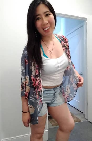 Person smiling, wearing a floral kimono implicit    a achromatic  apical  and denim shorts. They’re lasting  indoors, with a doorway disposable   successful  the background
