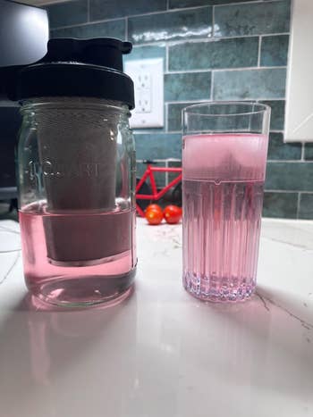 Glass and pitcher of pink drink on a kitchen counter with a backdrop of green tiles and tomatoes