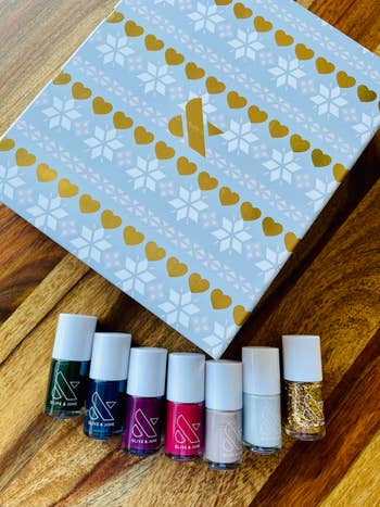 A blue box with six nail polish bottle on top in dark green, dark blue, maroon, pink, beige, light grey, and gold sparkle