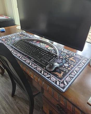 Desk setup with a large, decorative floral-patterned mouse pad, keyboard, and mouse in front of a monitor on a wooden table