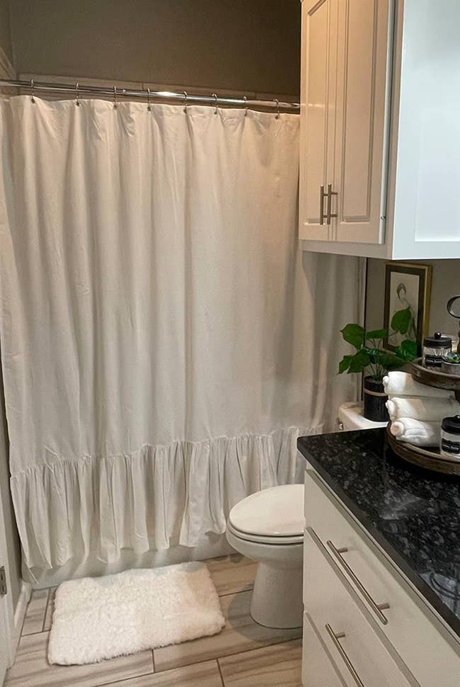 A modern bathroom featuring a ruffled shower curtain, white rug, black granite countertop, white cabinetry, and a plant on the counter