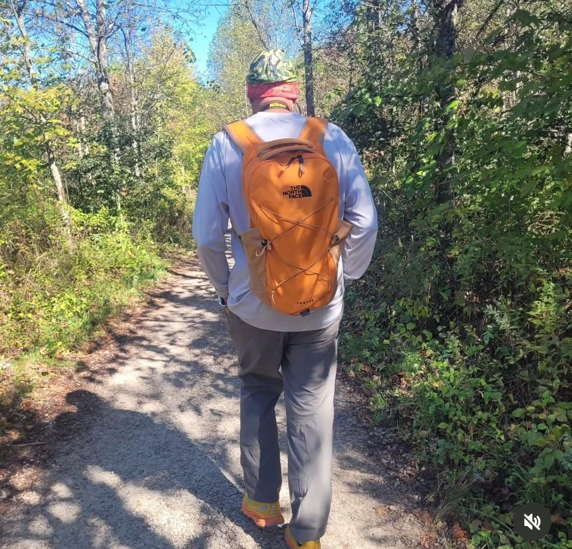 Person hiking connected  a wooded trail, wearing a backpack and casual outdoor clothing, surrounded by trees and foliage