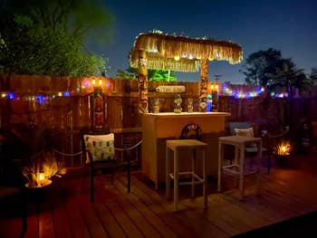 Outdoor tiki barroom  astatine  nighttime  with bamboo stools, tropical decor, drawstring  lights, and a writer  hut roof. Palm trees and plants situation   the woody  platform  area