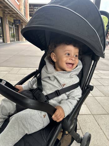 A smiling baby sits in a stroller wearing a grey hoodie and sweatpants, outdoors in a shopping area visible with store signage in the background