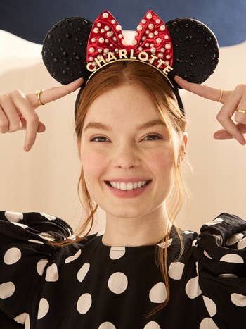 Person wearing a polka dot formal  and a Minnie Mouse ears headband with a reddish  bow, smiling and pointing to the headband