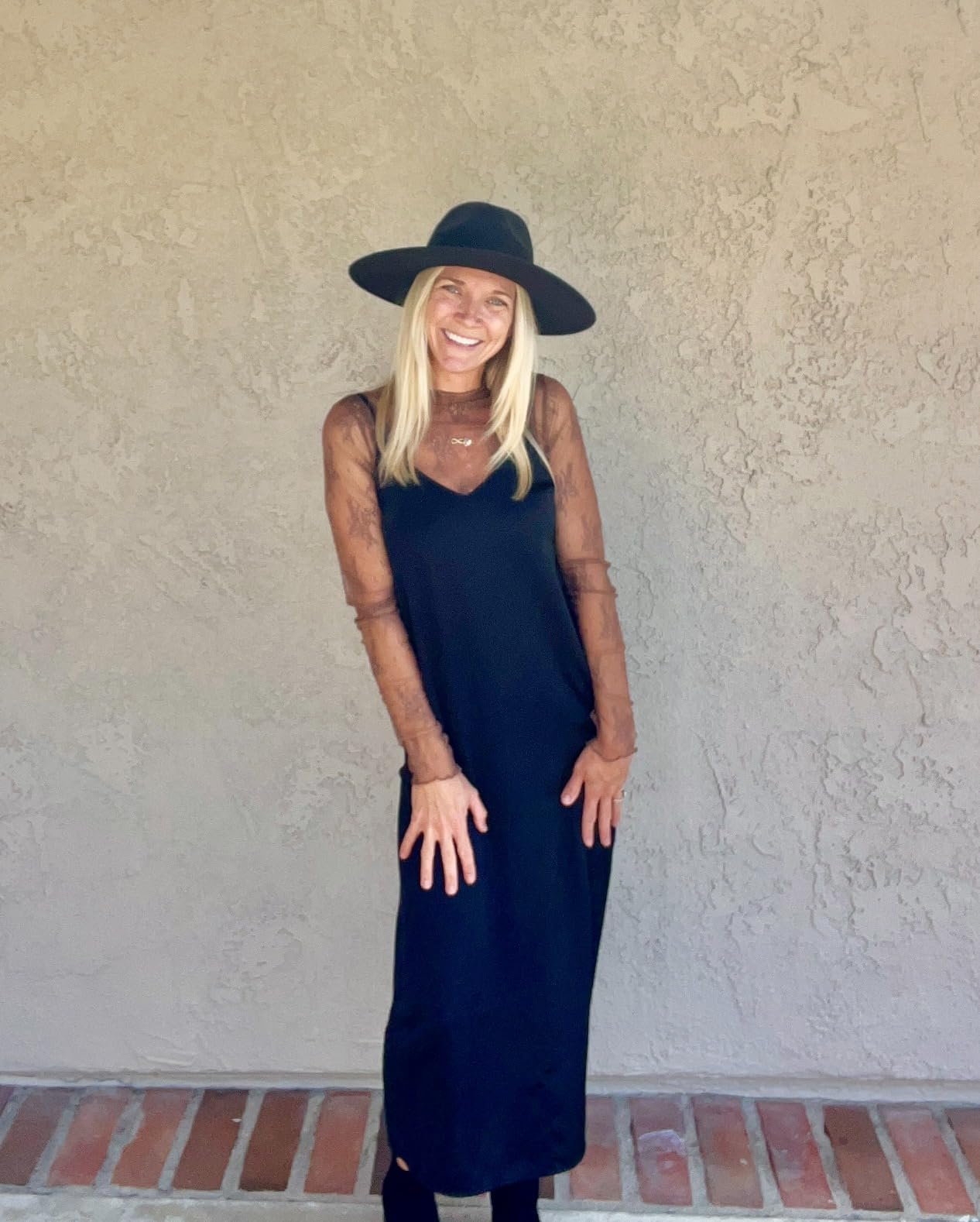 A reviewer smiling in wide-brim hat, sheer-sleeve dress, posed against a textured wall, in a shopping-focused article