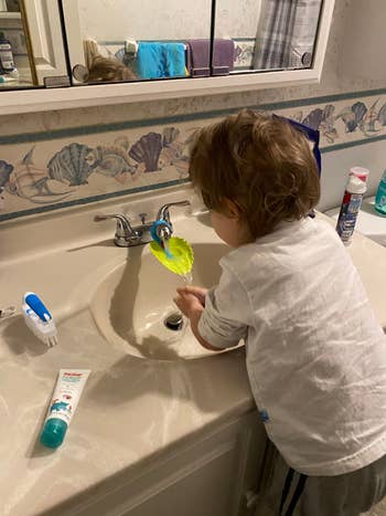 A young child is washing a green plastic leaf under the faucet in a bathroom. Various toiletries are visible on the counter, including toothpaste and a toothbrush