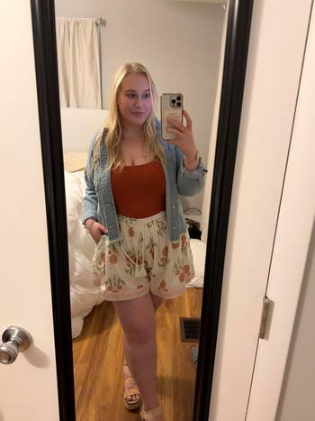 A person takes a mirror selfie wearing a denim jacket, a tank top, and floral shorts in a room with white bedding