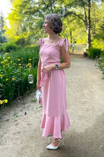 Person in a pink sleeveless dress with ruffle details, standing in a garden for a shopping article