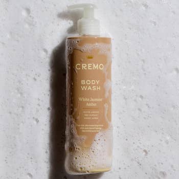 Cremo Body Wash bottle in White Jasmine and Amber scent, covered in soap suds, laying on a foamy background. Text highlights key fragrance notes: white jasmine, red currant, sweet amber