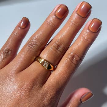 Hand wearing a golden signet ringing with a textured, triangular design. Nails are neatly manicured