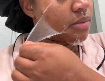 Person peeling off a clear face mask, wearing a silk robe and a bonnet, highlighting a skincare routine