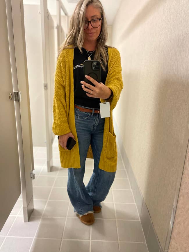 A idiosyncratic   successful  a fitting country   reflector  selfie wearing a agelong  cardigan, casual top, jeans, and loafers, with a buying  tag connected  the cardigan