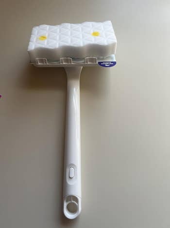Cleaning tool with a long handle and a textured sponge head, designed for scrubbing or dusting surfaces