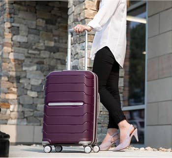 Person successful stylish outfit walking with a hard-shell suitcase connected wheels adjacent a modern gathering exterior