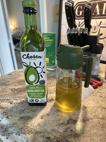 A bottle of avocado oil stands beside an oil dispenser on a kitchen counter, with a knife block and cleaning supplies in the background