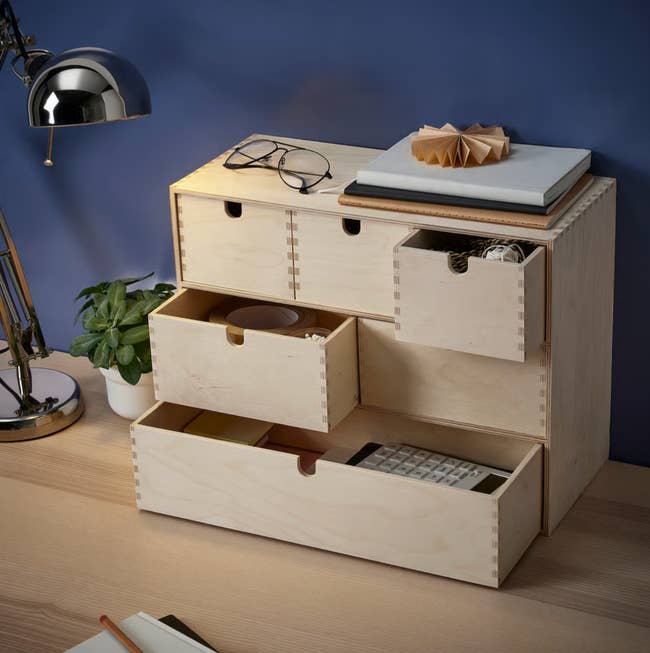 Desktop organizer with wooden drawers, a plant, glasses, lamp, and a notebook on top, suitable for efficient workspace storage