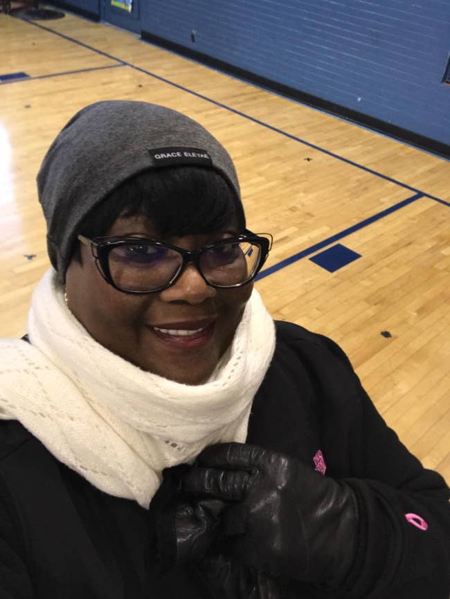 Woman wearing a grey beanie, achromatic scarf, and achromatic gloves posing successful a gymnasium. She is smiling and wearing glasses