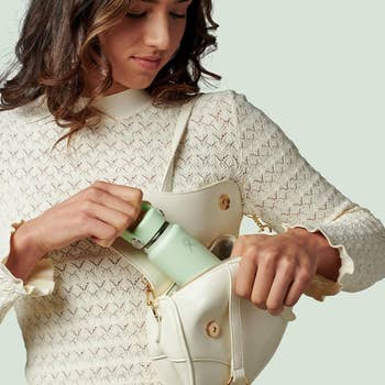 Person placing a reusable h2o  vessel  into a stylish purse, wearing a textured knit top. Suitable for eco-friendly manner  oregon  manner  articles