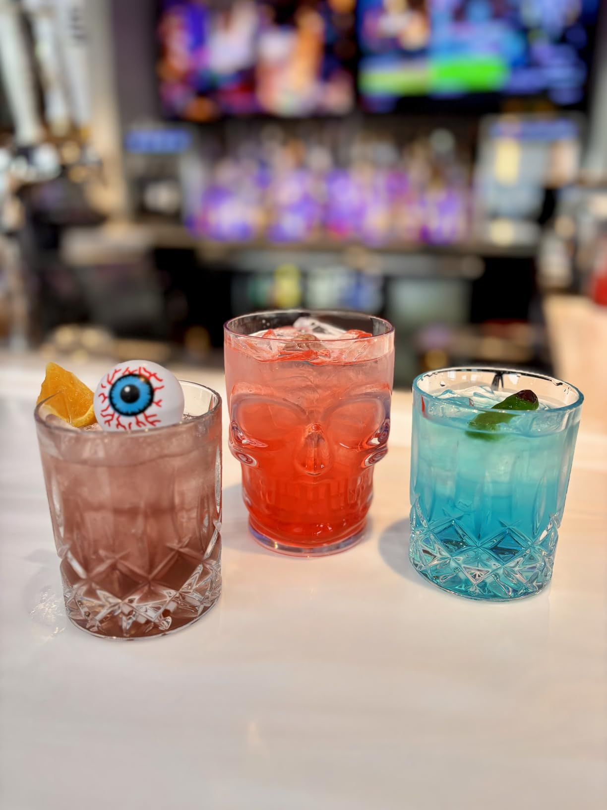  a skull glass, a garnished pinkish  portion  with an eyeball decoration, and a bluish  cocktail with a lime slice