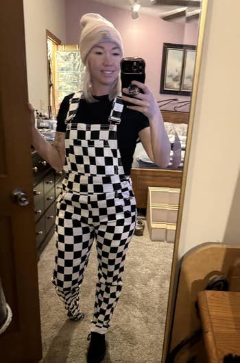 Person successful checkered overalls, achromatic tee, and beanie, taking a reflector selfie successful a casual chamber setting