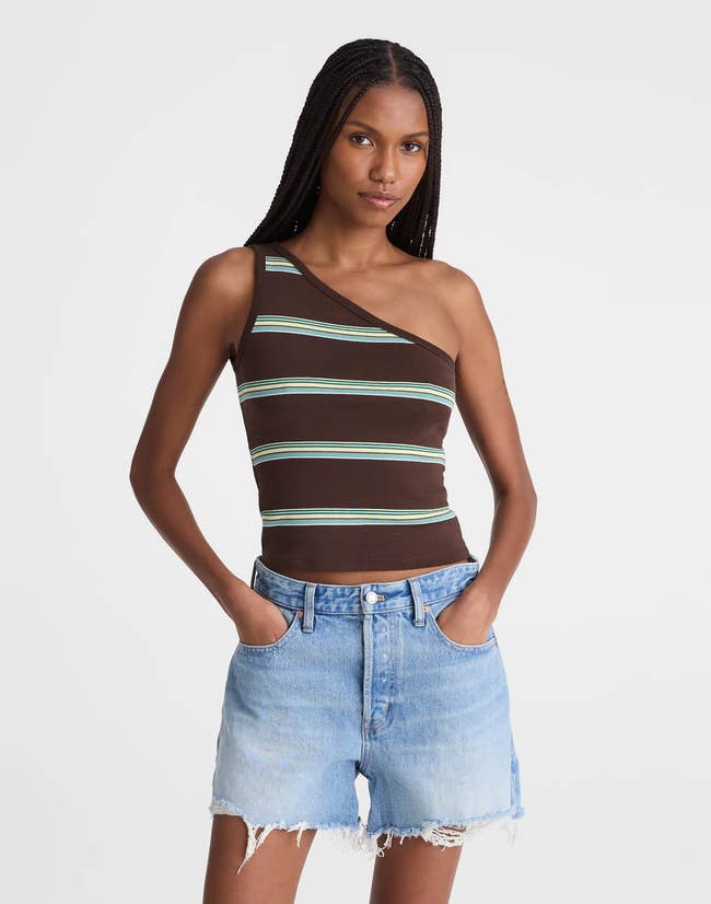 A model wears a stylish one-shoulder striped tank paired with frayed denim shorts, hands in pockets, suitable for a casual look