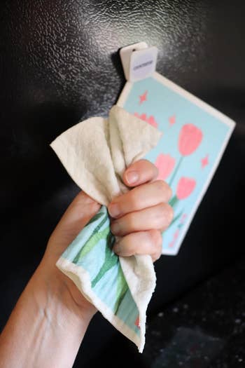Hand holding a room cloth with tulip plan successful beforehand of a fridge; a floral paper is magnetically attached above