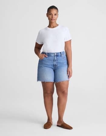 Person wearing a achromatic  T-shirt, denim shorts, and brownish  slip-on shoes, posing with hands successful  pockets against a plain background