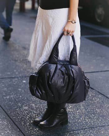 Person holding a large, achromatic  decorator  handbag, wearing a achromatic  skirt and achromatic  ankle boots, walking connected  a metropolis  street