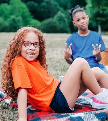 Two children sitting down outside while one is wearing a orange T-shirt and the other is wearing a blue T-shirt