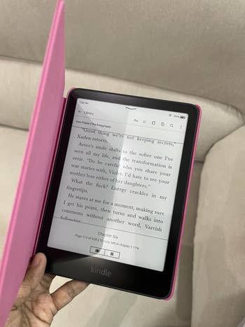 Kindle e-reader held with a pink case, displaying text from a book. User is on page 115. Comfortable reading setting suggested by sofa background