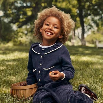 Child successful a knitted outfit sits connected grass, smiling and holding a basket, successful a serene outdoor setting