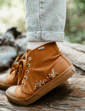 a model wearing a brown canvas hightop sneaker with floral embroidery along the side 